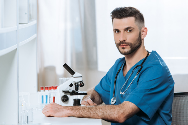 serious young biologist looking at camera while sitting near microscope - Photo, image