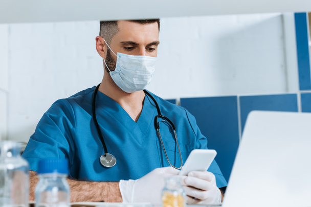 Laptop 'un yanında akıllı telefon kullanan tıbbi maskeli ve lateks eldivenli genç bir doktor. - Fotoğraf, Görsel
