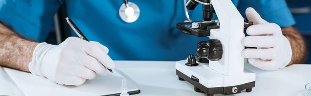 μερική άποψη του βιολόγου σε γάντια λατέξ γράφοντας στο σημειωματάριο κοντά στο μικροσκόπιο, πανοραμική λήψη - Φωτογραφία, εικόνα