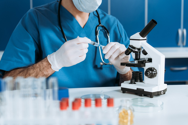 Tıbbi maskeli bir biyolog ve mikroskobun yanında şırınga tutan lateks eldivenler. - Fotoğraf, Görsel
