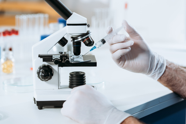 Ausgeschnittene Ansicht eines Biologen in Latexhandschuhen, der Spritze in der Nähe des Mikroskops hält - Foto, Bild