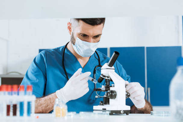 Tıbbi maskeli biyolog ve mikroskobun yanında şırınga tutan lateks eldivenli seçici odak.  - Fotoğraf, Görsel