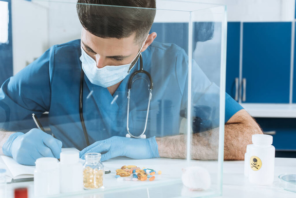 fiatal állatorvos orvosi maszk és késlelteti kesztyű írás közelében tartályok gyógyszerekkel és fehér egér üvegdobozban - Fotó, kép