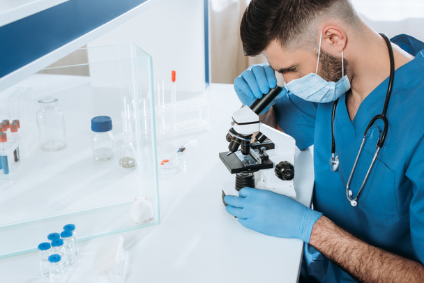 jovem biólogo em máscara médica e luvas tardias fazendo análise com microscópio perto do mouse branco em caixa de vidro e recipientes com medicamentos
 - Foto, Imagem