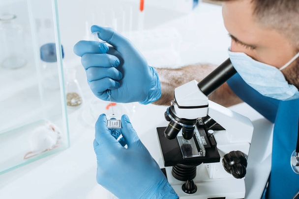 jonge bioloog in medisch masker en laat handschoenen met spuit terwijl hij een analyse maakt met een microscoop in de buurt van witte muis in een glazen doos - Foto, afbeelding