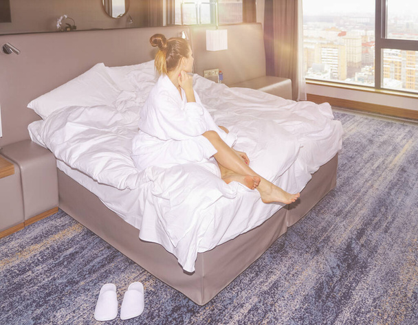 Young beautiful woman woke up and is sitting in the white bathrobe in her bed on the hotel window background at the yellow sunrise time. - Photo, Image