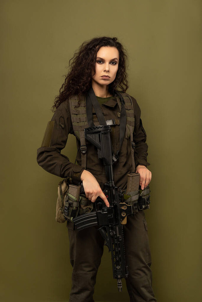 Mulher militar em uniforme verde com um colete descarregando com uma metralhadora em suas mãos em um fundo cáqui
 - Foto, Imagem