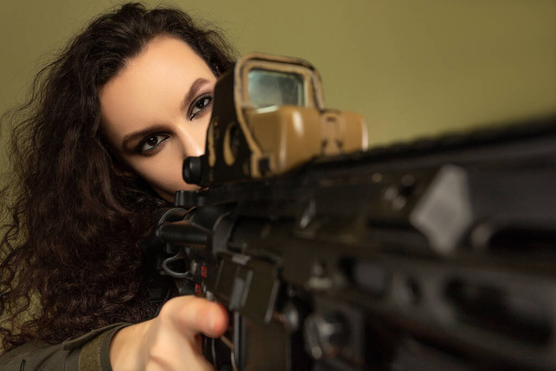 Militaire vrouw in groen uniform met een vest uitladen met een machinegeweer in haar handen op een kaki achtergrond. Een soldaat richt zich op de tactische aanblik van een geweer... een aanblik van een wapen. - Foto, afbeelding