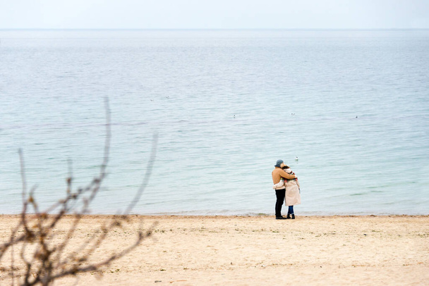 Happy loving couple embracing wearing warm clothes standing on ocean coast. Cold season, sea shore, empty beach in autumn or winter - Fotoğraf, Görsel
