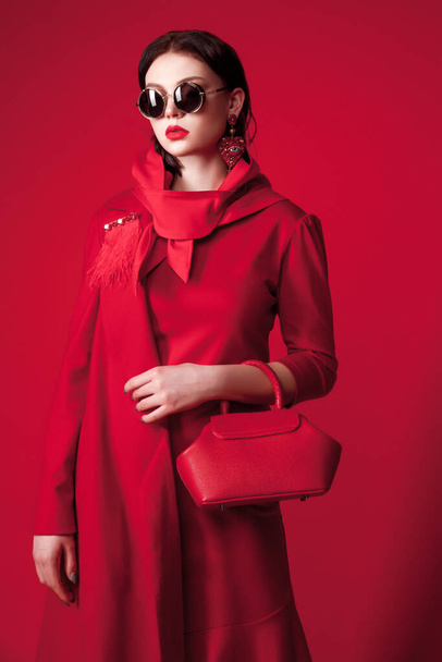 young beautiful woman in red trench coat holding her clutch. lady in sunglasses on red background. trendy outfit. creative fashion concept. monochrome outfit - Photo, Image