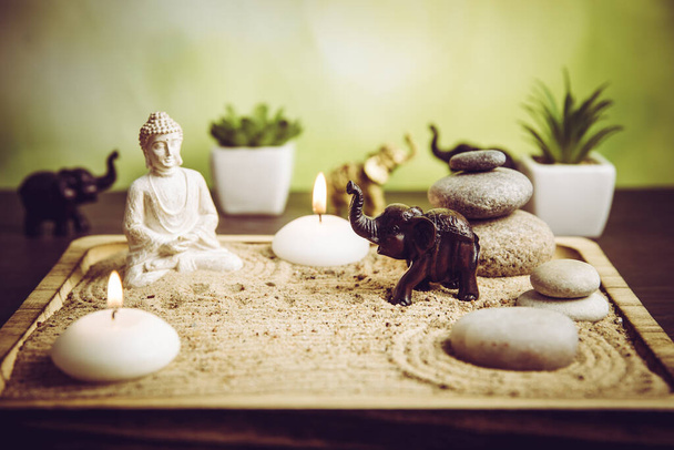 Selective focus on brown elephant figurine, miniature desk zen sandbox with Buddha figure sit in Lotus position, stacked zen sea stones, spa candles burning against green bokeh studio background. - Foto, afbeelding