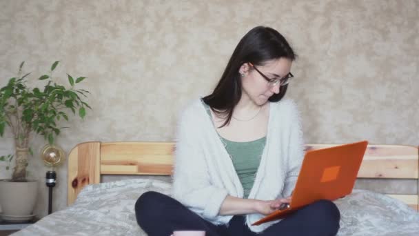 Young attractive girl is actively browsing the news feed on a laptop while sitting on a bed at home. Young Cute girl is working on a laptop while sitting on a bed. Working at home. - Materiał filmowy, wideo