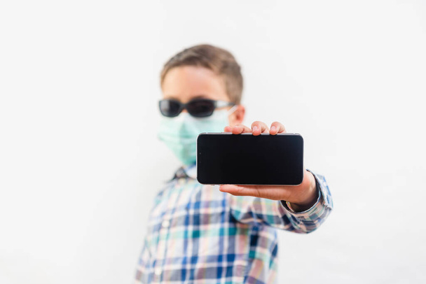 Young boy wearing disposable mask and sunglasses holding mobile phone over white background - Valokuva, kuva