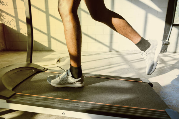 ランニングマシンやサンレイ付きフィットネスジムでトレッドミルで実行されているフィットネスマンの足の部分で下体。暖かい音色だ。健康と運動活動の概念.ワークアウトと力トレーニングのテーマ. - 写真・画像