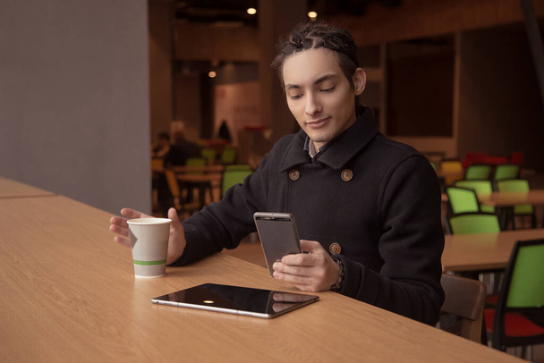 Mladý muž sedí v kavárně s chytrým telefonem a pije kávu. Hezký brunetka chlap s dredy, v kabátě. - Fotografie, Obrázek