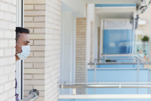 closeup of a young caucasian man wearing a surgical mask leaning out the window of his house or of a hotel room - Photo, image