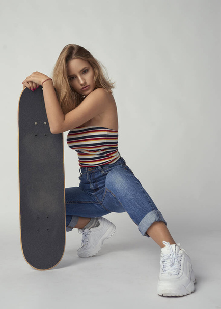 молодая и спортивная девушка со скейтбордом в джинсах и короткой футболке, красивой и здоровой кожей, натуральными светлыми волосами, пустым фоном, нежным макияжем
 - Фото, изображение