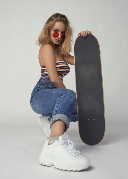 uma menina jovem e atlética com um skate em jeans e uma camiseta curta, pele bonita e saudável, cabelo loiro natural, fundo vazio, maquiagem delicada
 - Foto, Imagem