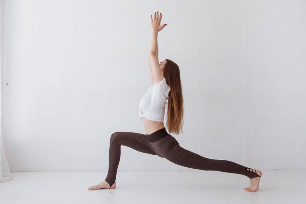Side view portrait of beautiful young woman wearing white tank top working out against white wall, doing yoga or pilates exercise. Standing in Warrior one pose, Virabhadrasana. Full length - Φωτογραφία, εικόνα