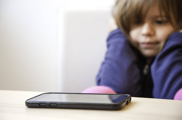 Focus selettivo di uno smartphone sul tavolo in legno. Sullo sfondo una bambina guarda lo smartphone, ma non vuole toccarlo. L'infanzia. Consapevolezza degli effetti tecnologici sui bambini
. - Foto, immagini