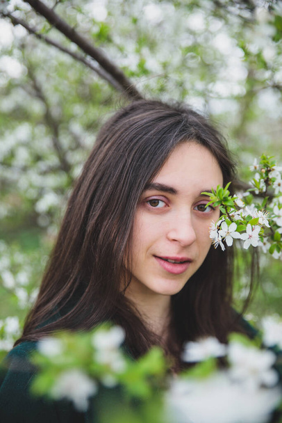 Young caucasian girl with dark hair in white t-shirt and green coat near beautiful white cherry blossom tree spring nature background - Fotó, kép