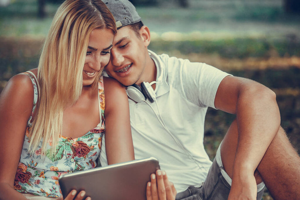 公園でピクニックをしながら、タブレットでオンラインでメディアコンテンツを見て幸せな若いカップルのショット.  - 写真・画像
