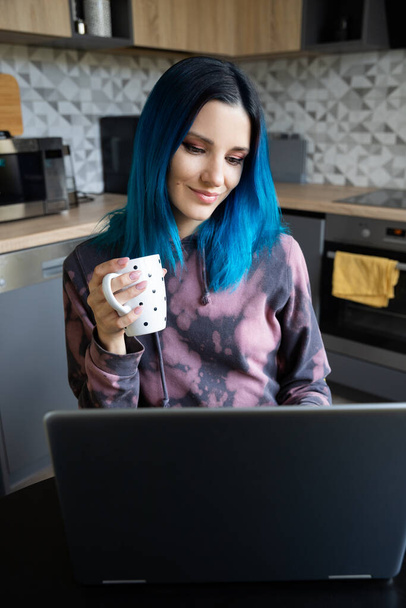 Joyeux jeune femme hipster avec les cheveux bleus se détendre avec ordinateur portable sur la cuisine. Succès souriant travail féminin du concept de la maison
. - Photo, image