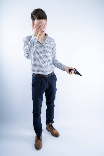 Young caucasian criminal man over gray background holding gun stressed with hand on head, shocked with shame and surprise face, angry and frustrated. Fear and upset for mistake. - 写真・画像