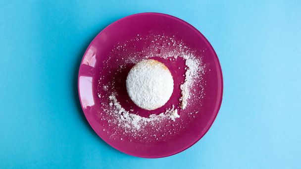 Sprinkle one cottage cheese with powdered sugar on a pink plate, on a blue background, top view. Dessert, a small cupcake. Food concept. white baked cookies with an airy texture. Copy space. - Photo, Image