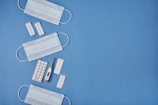 Medicine masks, electronic thermometer and pills blister packs arranged in a row vertically on the left side of image on blue background, top view with a lot of background - Foto, Bild