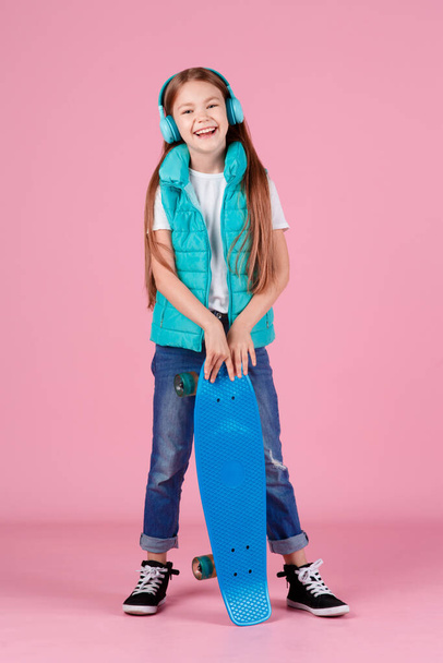 Beautiful little blonde kid girl 7-10 years old in jacket posing isolated on pastel pink background children portrait. Childhood lifestyle concept. Mock up copy space. Hold blue skateboard - Photo, image