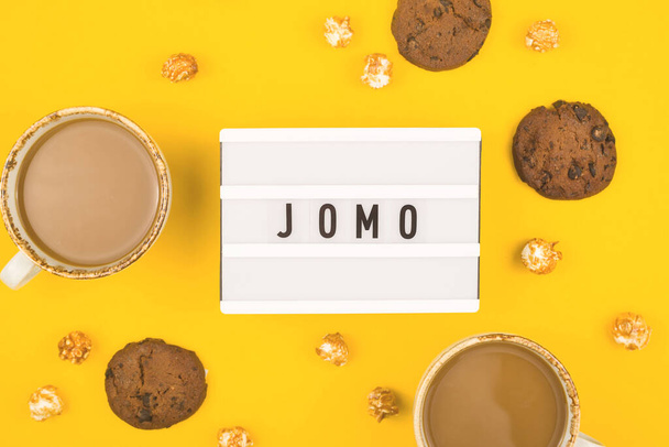 Abbreviation word JOMO written on a decorative board on a bright yellow background. The concept of relaxation from information and gadgets. Top view, flat lay. - Photo, Image
