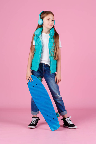 Beautiful little blonde kid girl 7-10 years old in jacket posing isolated on pastel pink background children portrait. Childhood lifestyle concept. Mock up copy space. Hold blue skateboard - Фото, изображение