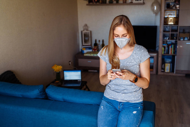 Schöne Mädchen liest Nachrichten über die Coronavirus-Epidemie am Telefon und ruft ihre Freunde an. Sie bleibt allein zu Hause und trägt Atemschutzmasken, während sie aus der Ferne arbeitet. - Foto, Bild