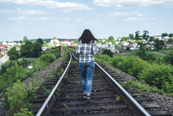 Jonge vrouw in jeans en shirt loopt op een zonnige dag langs het spoor - Foto, afbeelding