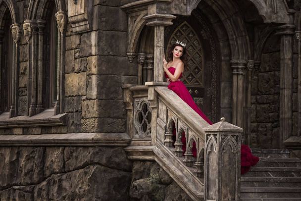 A beautiful woman, a queen in a red dress, stands on the background of a medieval, Gothic door on the stairs with an arch, silver caron with crystals. The Princess at the Castle - Fotó, kép