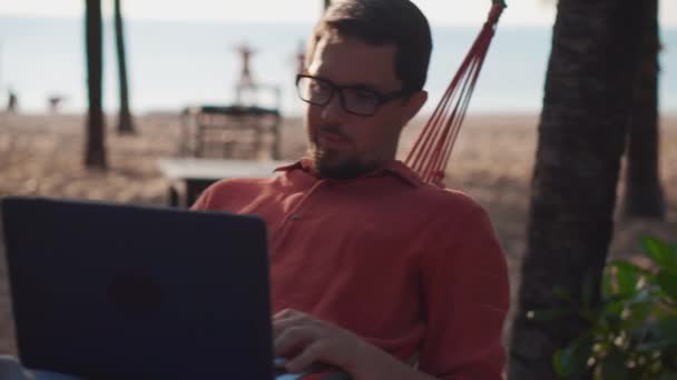 Muž relaxuje s laptopem v houpací síti na pláži - Záběry, video