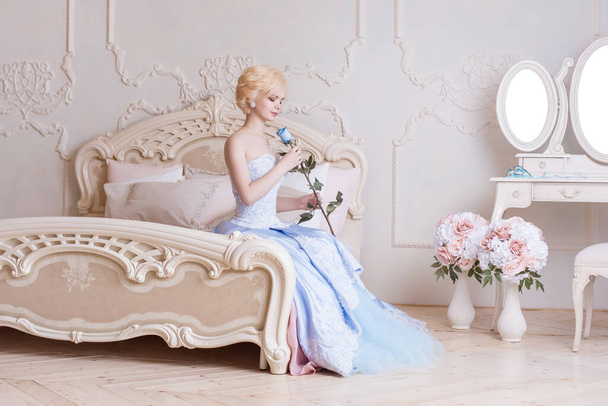 A beautiful, young blonde woman sits on a bed in a bedroom, dressed in a ball gown, and holds a blue rose in her hands. Princess in the Versailles, France. - Foto, imagen