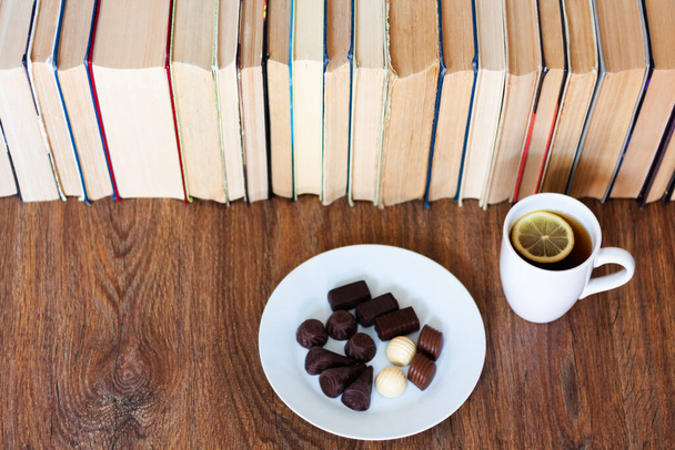 cup of tea and chocolate candies on white plate, stack of old book on wooden table, education concept background, many books piles with copy space for text - Photo, Image