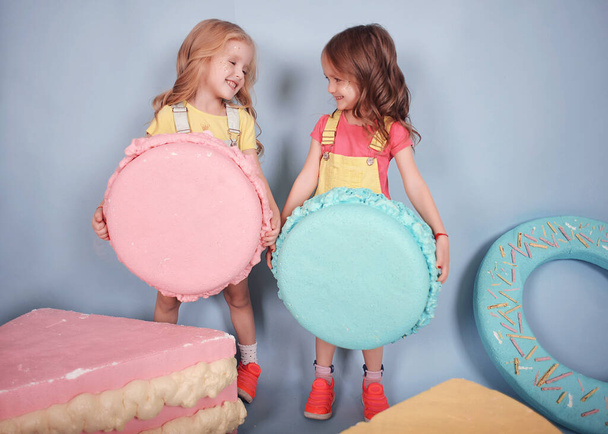 cute kids two little girls are having fun and playing on a blue background, they love sweets : cakes, donuts, sweets and macarons! Children have bright clothes, they laugh. hugging and loving, two twin sisters with a beautiful girl's mom - Photo, Image