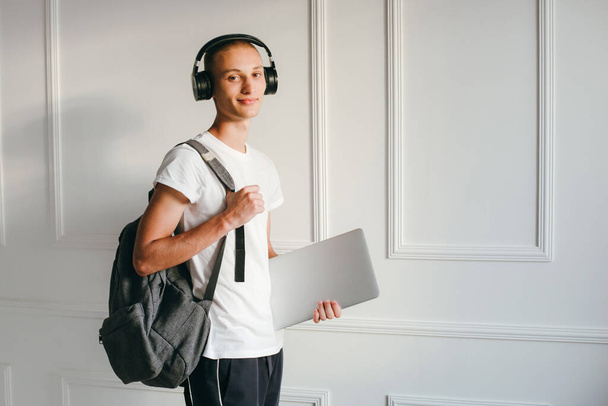 Adolescente menino estudante de pé segurando laptop e usando fones de ouvido sem fio e a mochila
 - Foto, Imagem