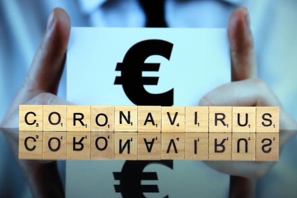 WROCLAW, POLAND - MARCH 30, 2020: The word CORONAVIRUS made of wooden letters, and man holding a business card with euro symbol in the background. Coronavirus (COVID-19) global disease 2020. - Fotoğraf, Görsel