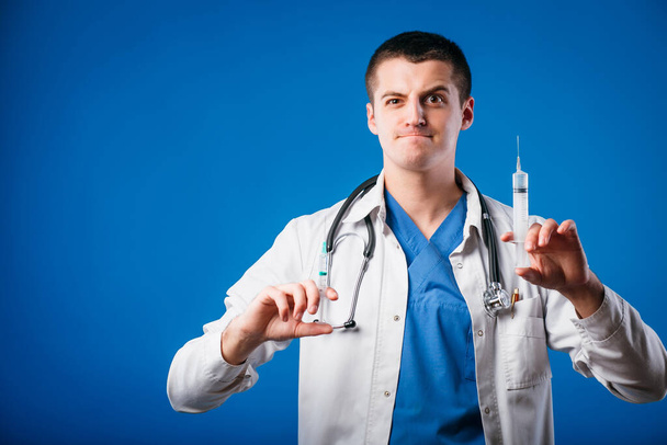 Портрет смешного эмоционального врача-мужчины с двумя шприцами, изолированными на синем фоне
 - Фото, изображение