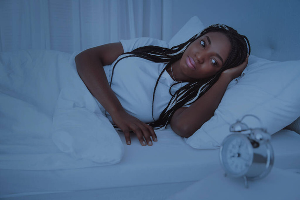 A beautiful African woman lies and bed, in a white bedroom and looks at the clock. Insomnia, rest at home. Healthy sleep. Nigeria, Africa. - Fotografie, Obrázek