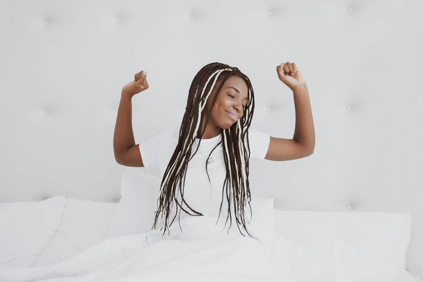 A beautiful African woman sits on a bed in a white bedroom and stretches. Good morning, vacation at home. The girl is wearing a T-shirt, pigtails on her head. Healthy sleep. Nigeria, Africa. - Foto, afbeelding