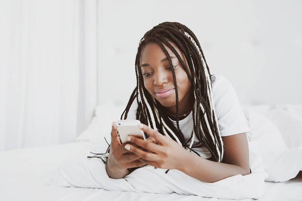 Eine schöne Afrikanerin liegt auf dem Bauch, auf dem Bett und schaut ins Telefon. Soziale Netzwerke checken, SMS verschicken. Das Mädchen trägt ein T-Shirt, Zöpfe auf dem Kopf. Porträt. - Foto, Bild