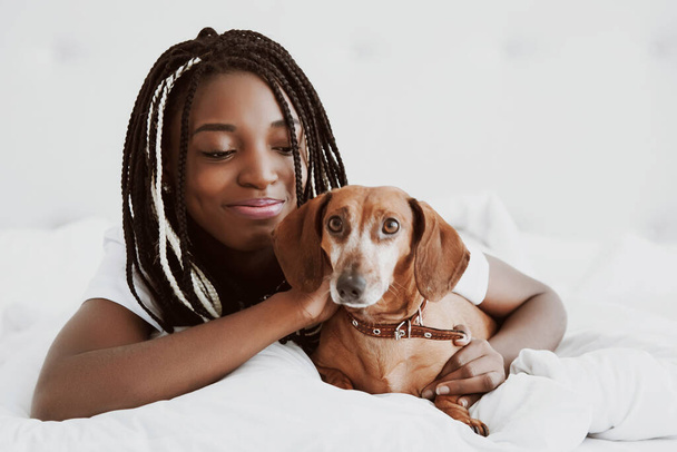 A beautiful African woman lies on her stomach, on a bed with a red dog dachshund, a domestic pet. Good morning. Portrait. Nigeria, Africa. - Фото, изображение