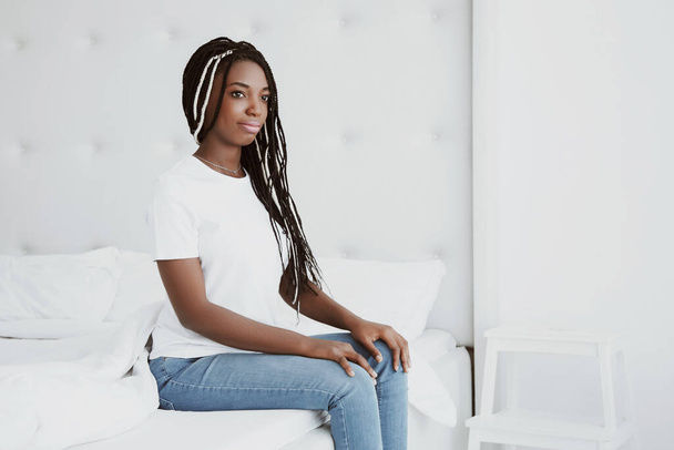 A beautiful African woman sits on the edge of a bed in a white bedroom. The girl is dressed in tight jeans and a T-shirt, top. Nigeria, Africa. Copy space, Your text here. - Фото, изображение