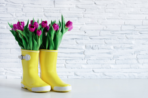 Yellow rain boots and purple tulips over white brick wall. Spring background. Copy space. - Foto, afbeelding