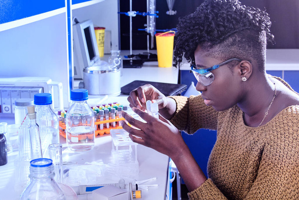 Nuori afrikkalainen naispuolinen teknologia tai tutkija suojalaseissa toimii biologisten tai lääketieteellisten nestemäisten näytteiden kanssa nykyaikaisessa tutkimuslaboratoriossa. Laboratorio-olosuhteiden määrittäminen lääketutkimusyksikössä - Valokuva, kuva